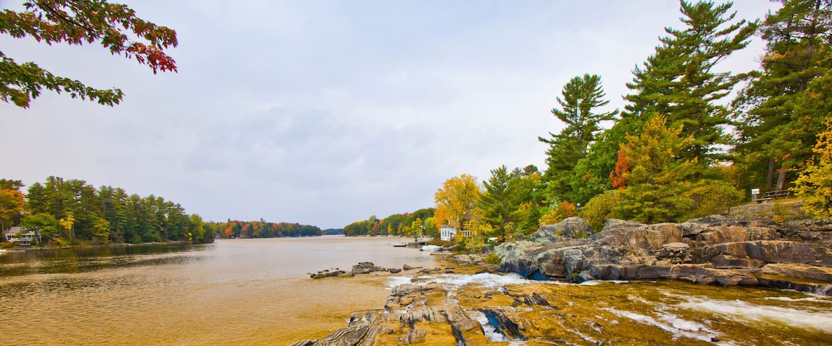 A river with rocky banks, surrounded by autumn-colored trees and a cloudy sky.