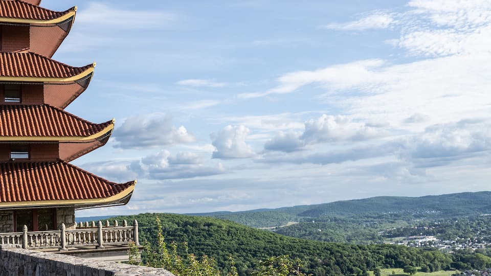 A traditional multi-tiered pagoda with a stone railing, set against a backdrop of lush green hills and a partly cloudy sky.
