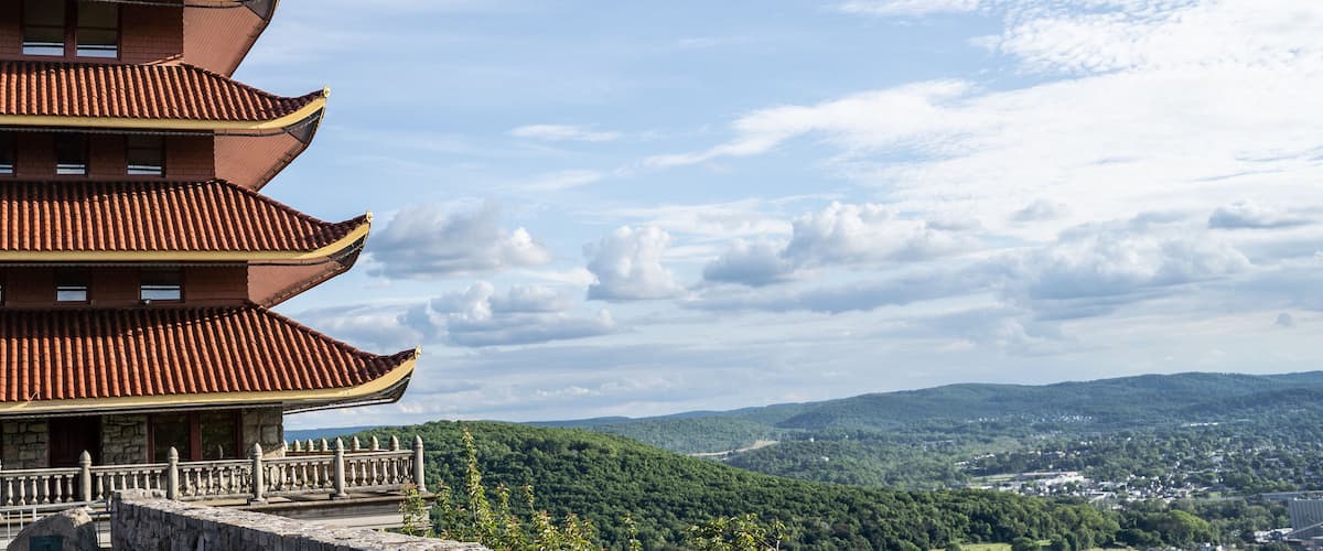 A traditional multi-tiered pagoda with a stone railing, set against a backdrop of lush green hills and a partly cloudy sky.