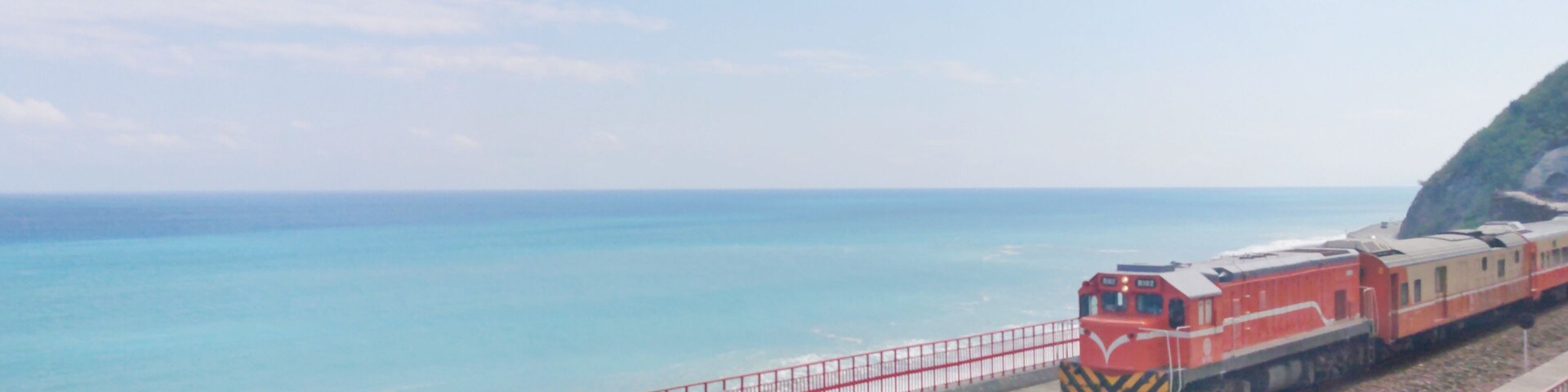 Um trem vermelho sobre trilhos perto de uma praia, com uma grade vermelha e um penhasco rochoso.