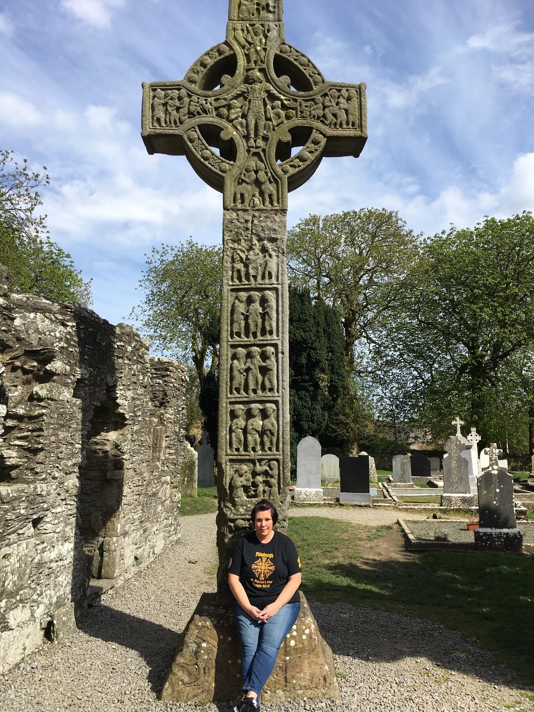 A large stone cross with intricate carvings, set in a cemetery with other tombstones and a stone wall in the background.