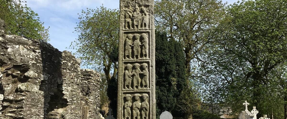 A large stone cross with intricate carvings, set in a cemetery with other tombstones and a stone wall in the background.