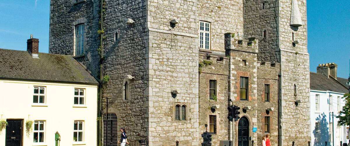 A historic stone castle with two towers, a white building with a black door, and a clear blue sky.