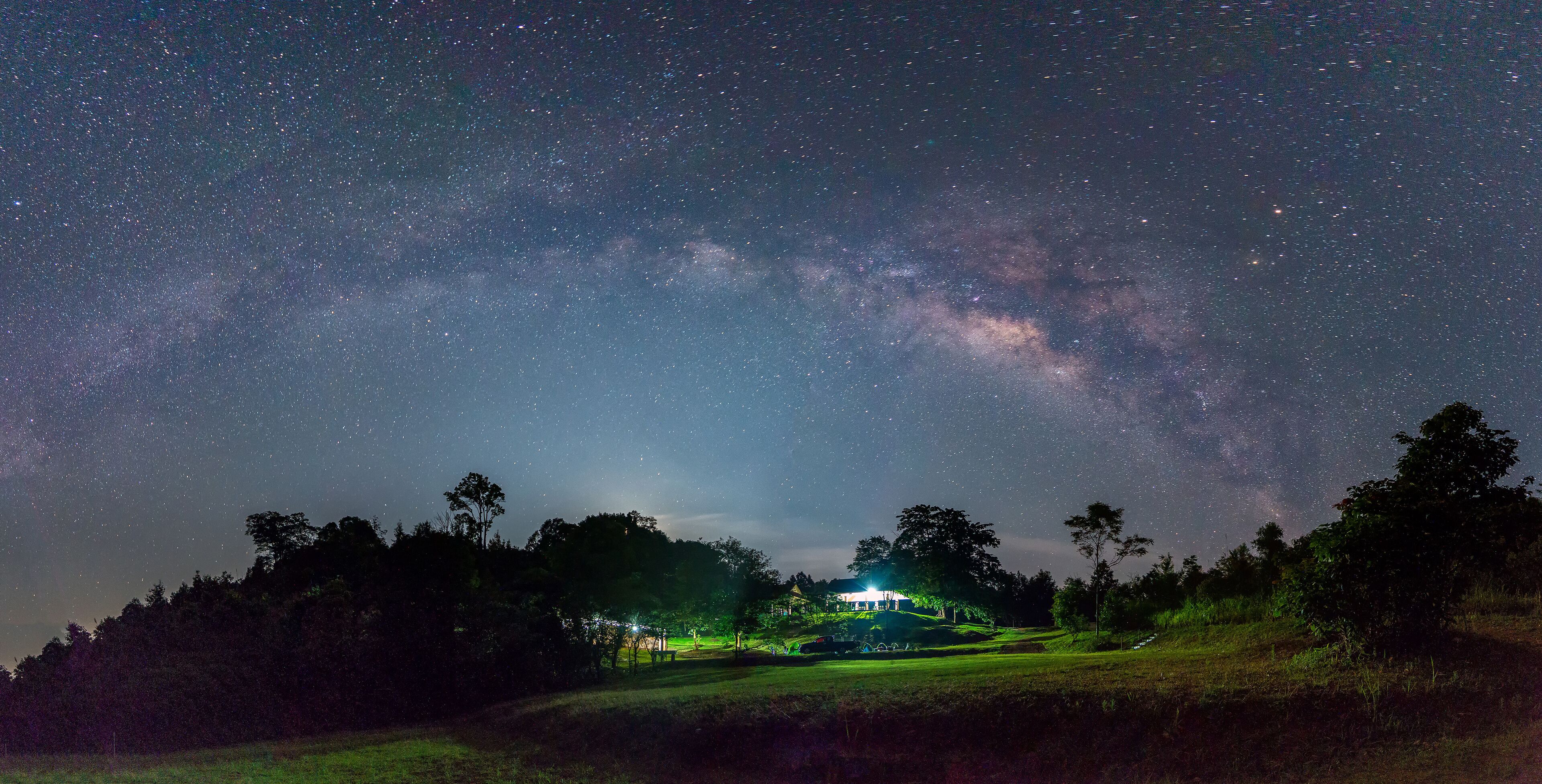 A starry night sky over a grassy field with a house and trees.