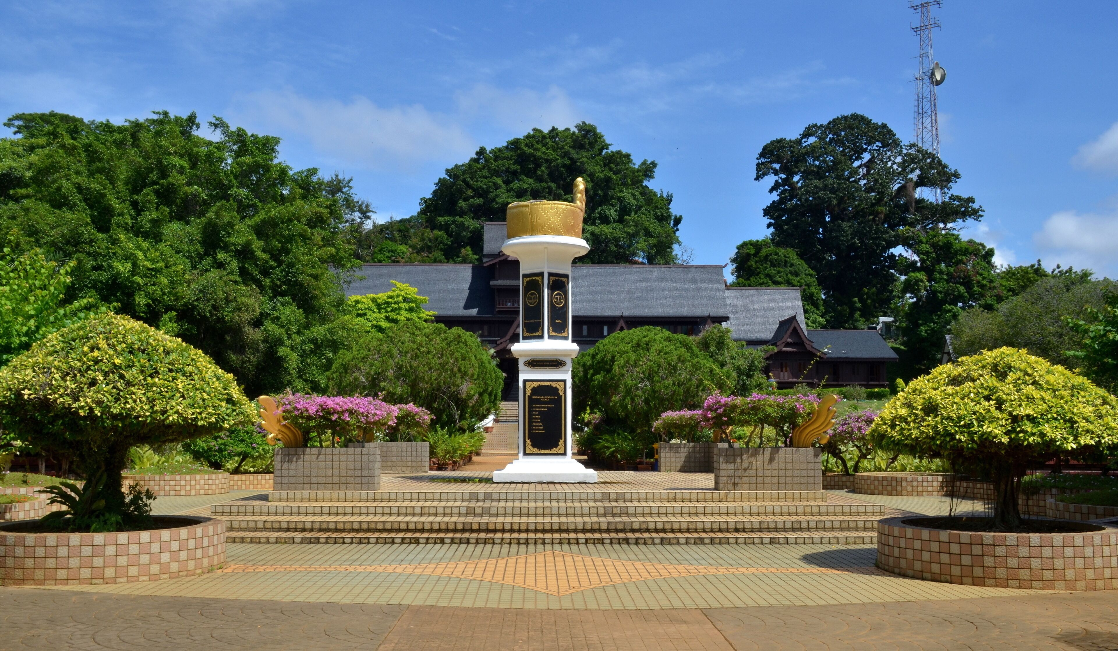 A white monument with a golden top, surrounded by well-manicured bushes and trees, set against a backdrop of a traditional building and a clear blue sky.