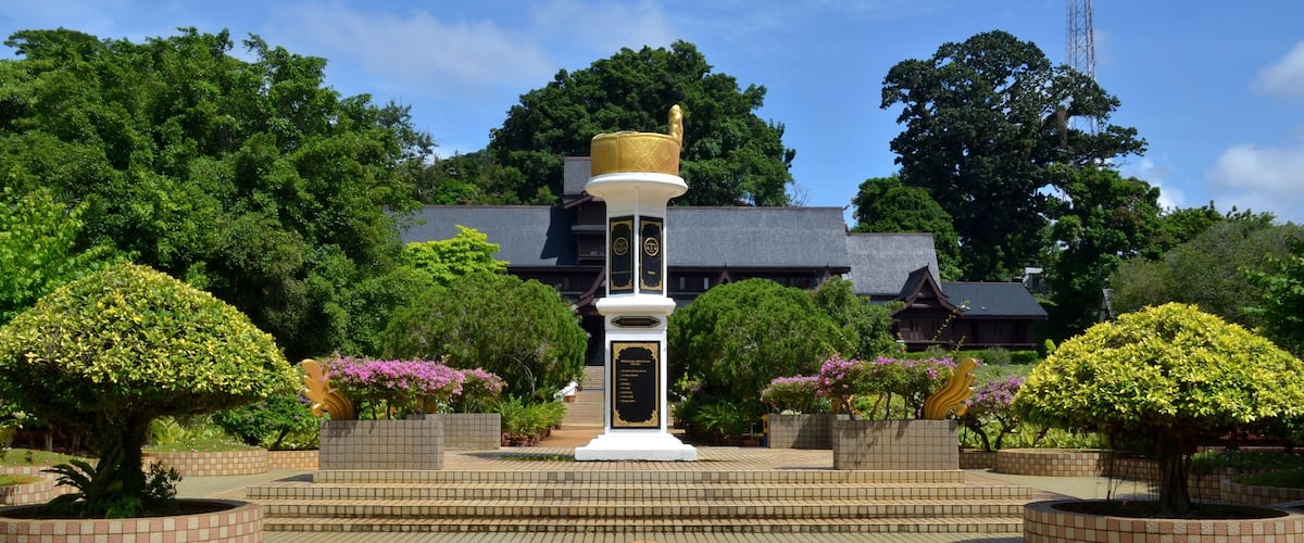 A white monument with a golden top, surrounded by well-manicured bushes and trees, set against a backdrop of a traditional building and a clear blue sky.