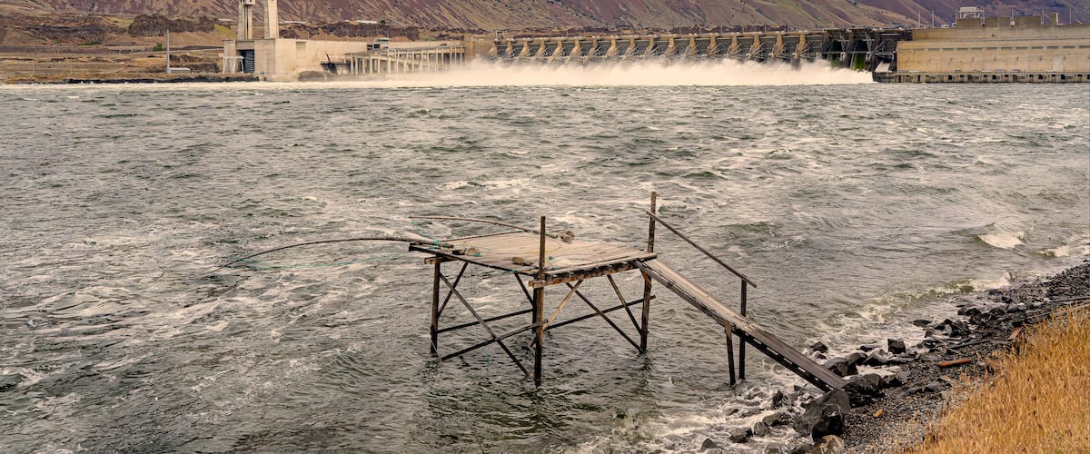 A dam with water flowing over it, a wooden pier, and a mountainous landscape during sunset.