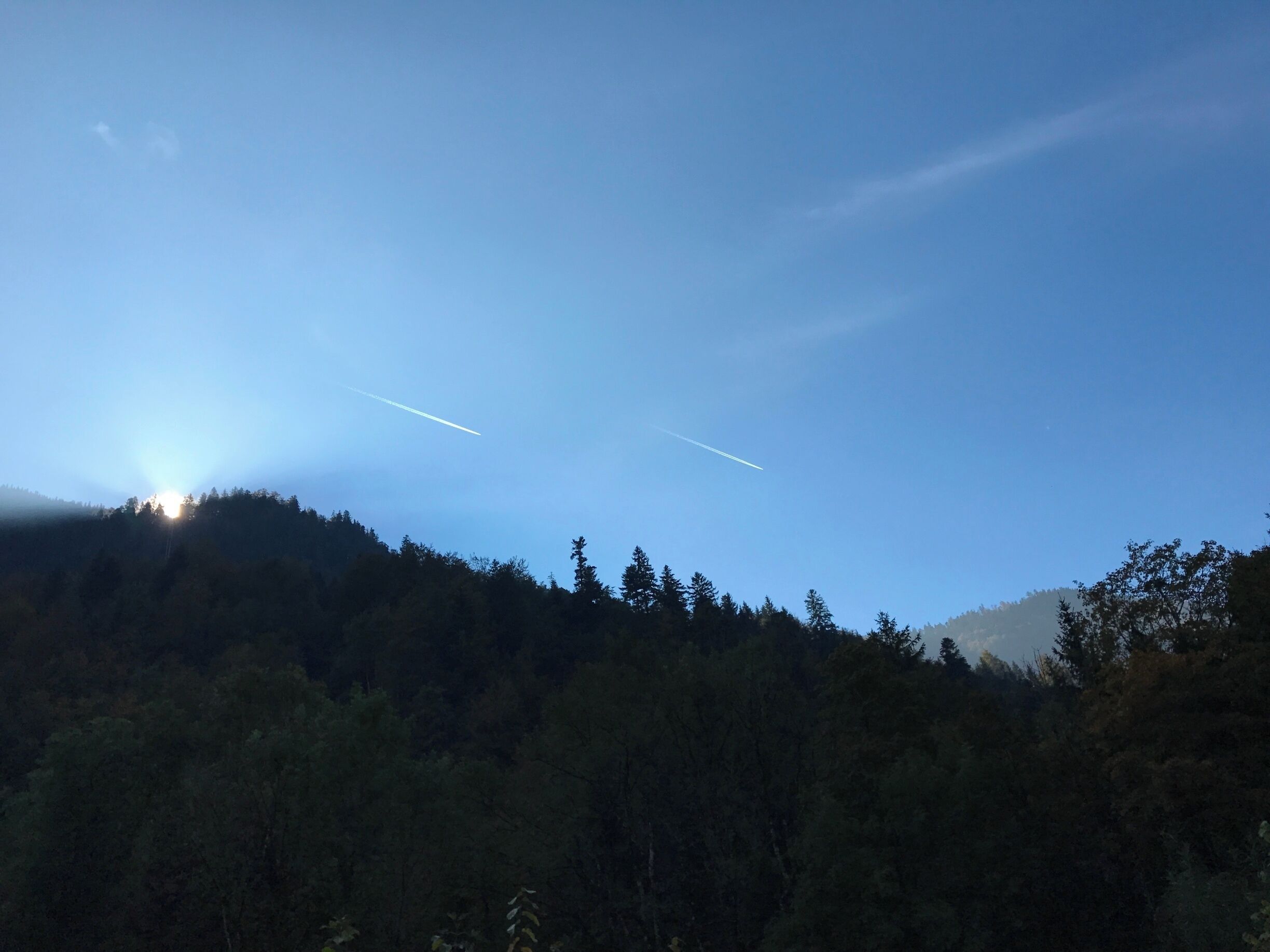 A clear sky with two contrails, a sun peeking through the trees, and a forested hillside.