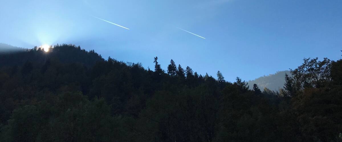 A clear sky with two contrails, a sun peeking through the trees, and a forested hillside.