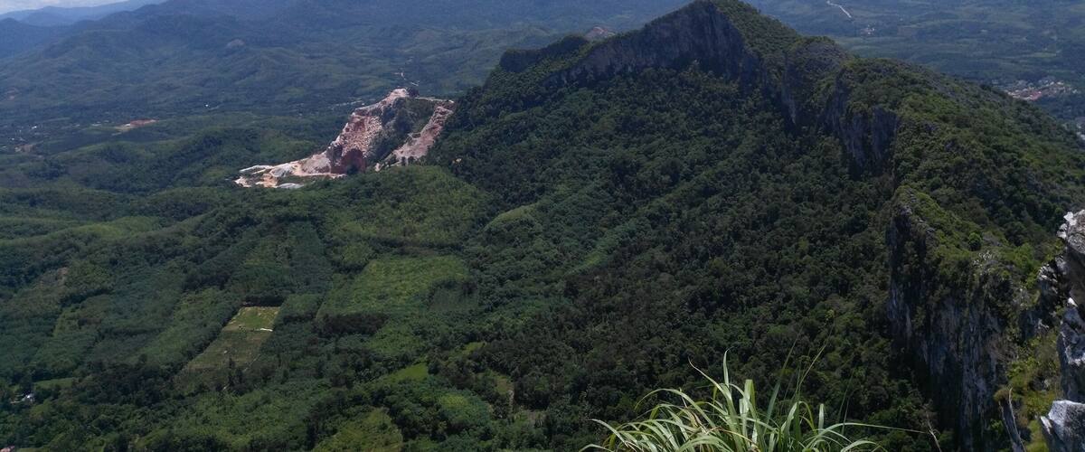 A mountainous landscape with dense green forests, a prominent rocky peak, and distant mountains under a partly cloudy sky.