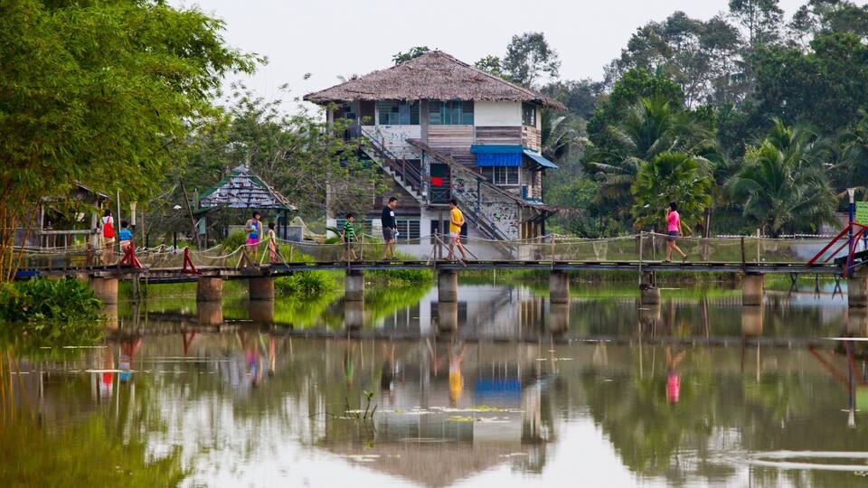 A thatched-roof building with a staircase, a bridge over a calm body of water, and lush greenery in the background.