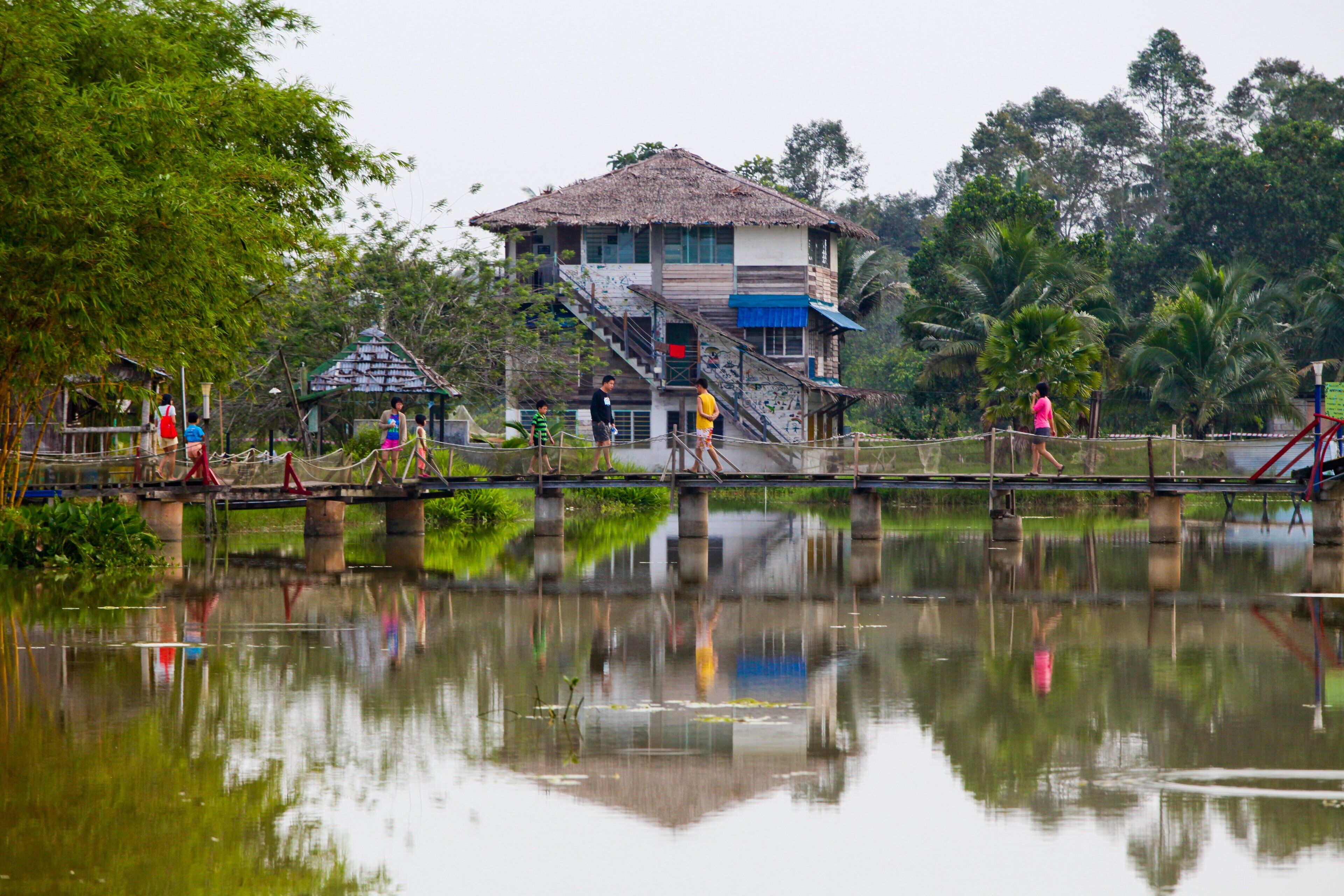 A thatched-roof building with a staircase, a bridge over a calm body of water, and lush greenery in the background.