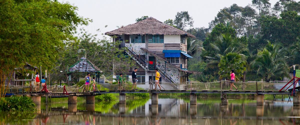 A thatched-roof building with a staircase, a bridge over a calm body of water, and lush greenery in the background.