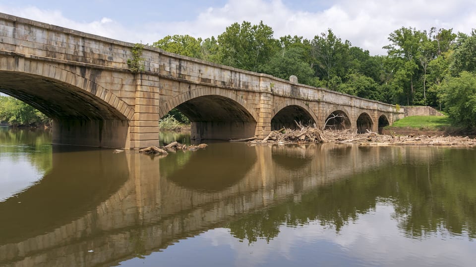 A stone bridge over a calm river with reflections, surrounded by trees.