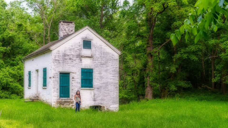 A white wooden house with green shutters in a lush green field surrounded by trees.