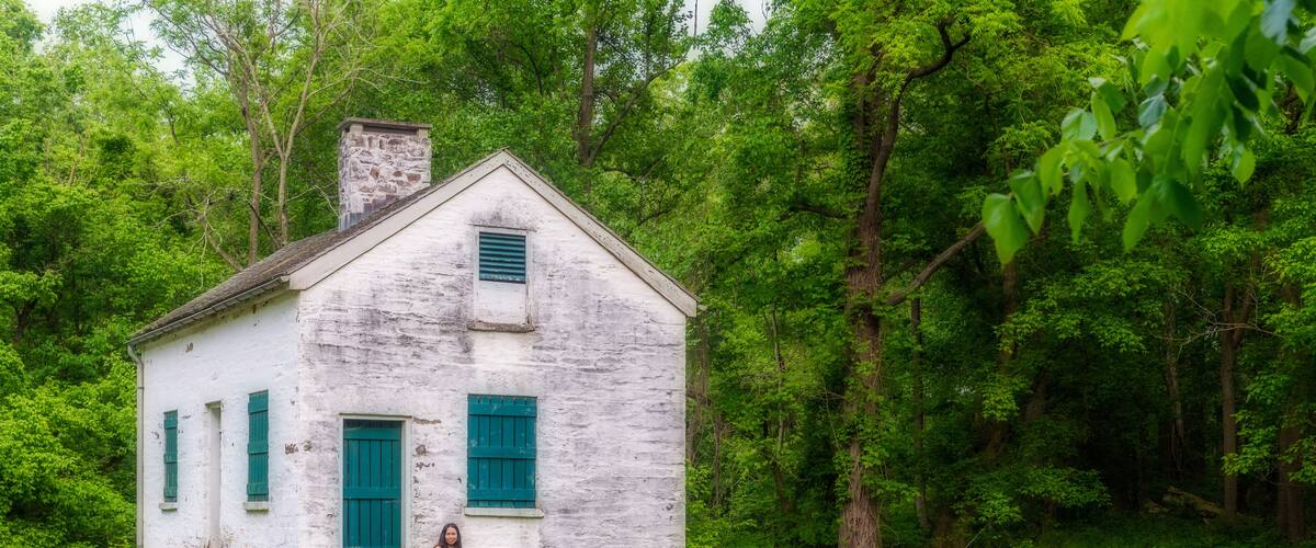 A white wooden house with green shutters in a lush green field surrounded by trees.