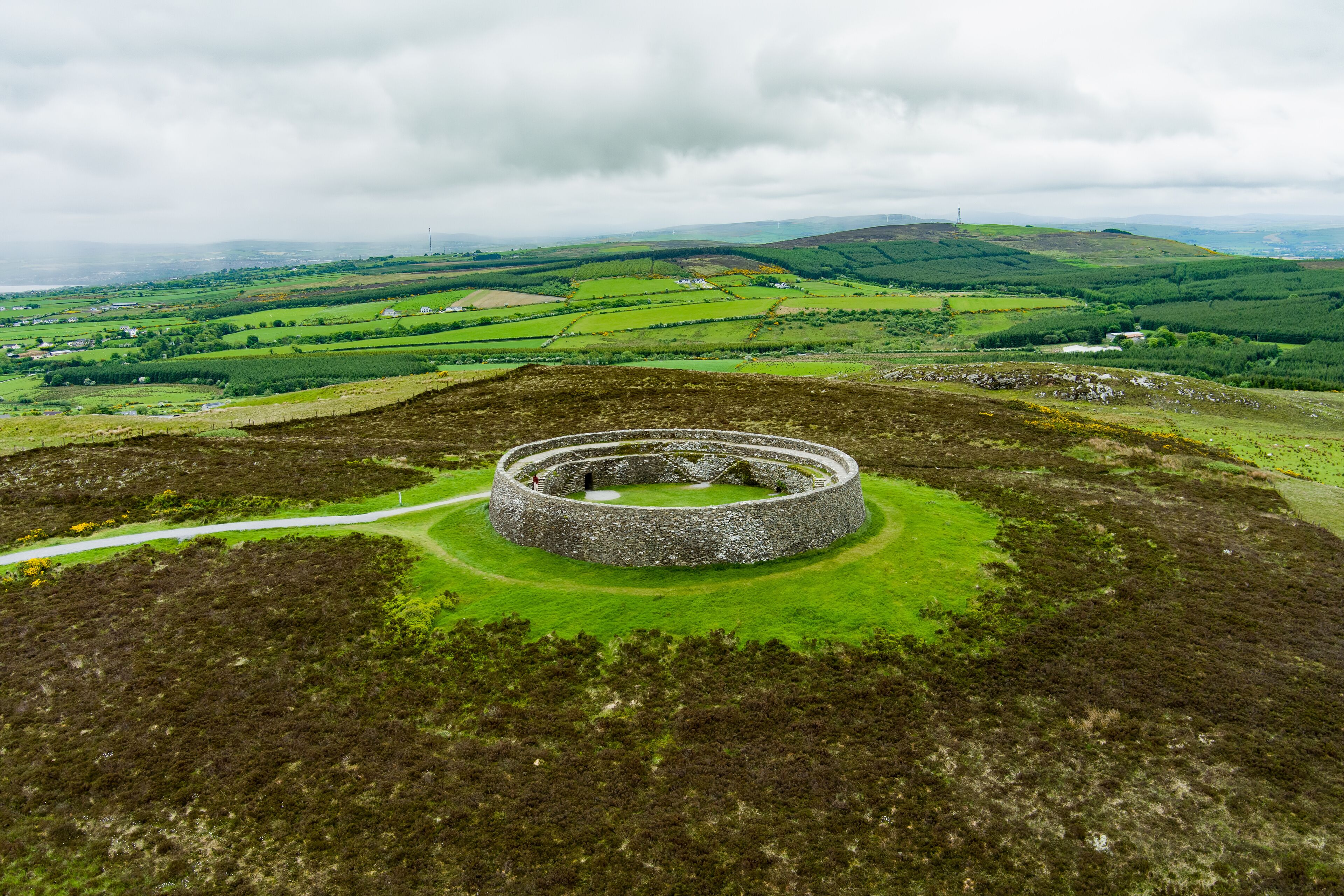 A circular stone structure in a green landscape with rolling hills and a cloudy sky.