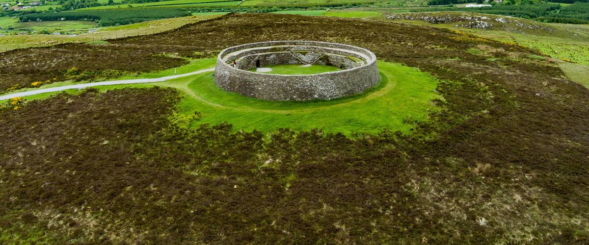 A circular stone structure in a green landscape with rolling hills and a cloudy sky.