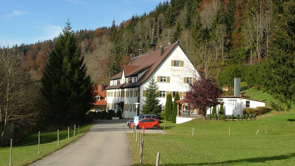 A white house with a red roof, a parked red car, and a forested hillside in the background.