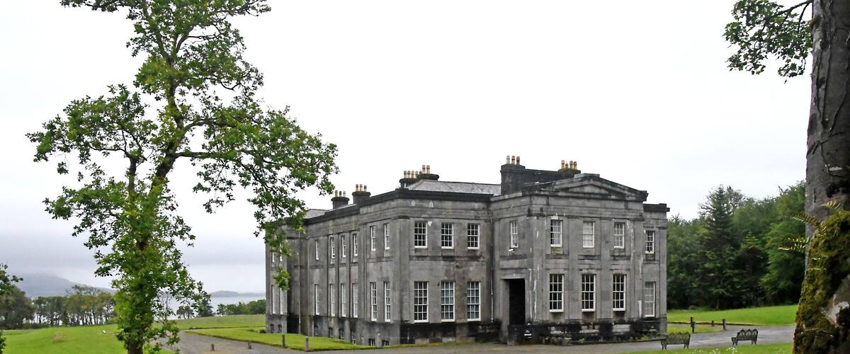 A large, historic stone building with a symmetrical facade, surrounded by lush green fields and a small body of water in the background.