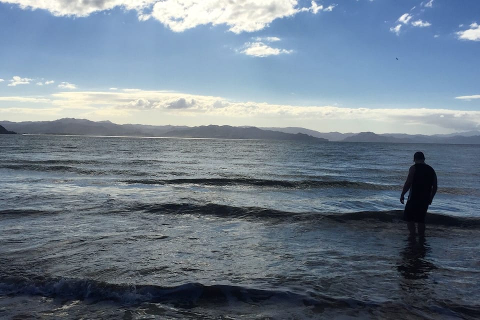 Una persona de pie en aguas poco profundas cerca de un cuerpo de agua, con montañas al fondo.