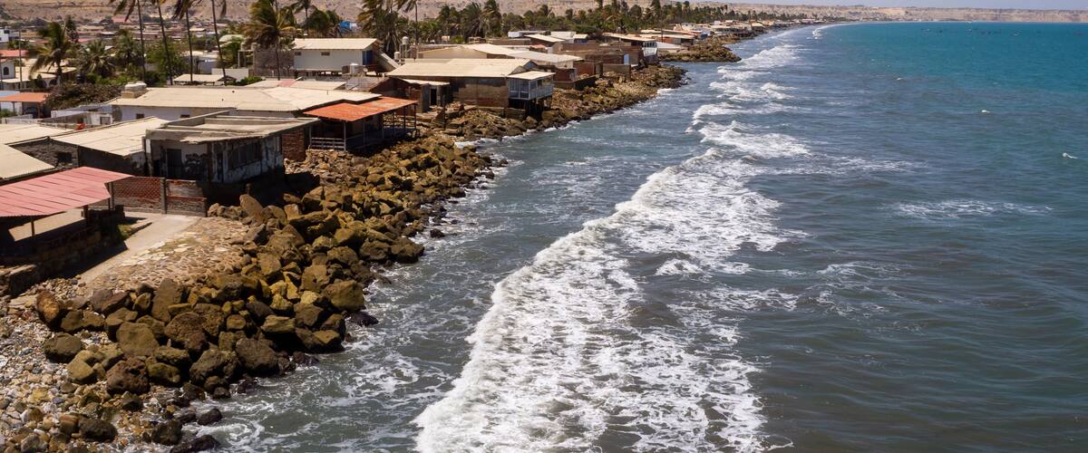 A coastal town with buildings along the shore, palm trees, and a rocky breakwater.