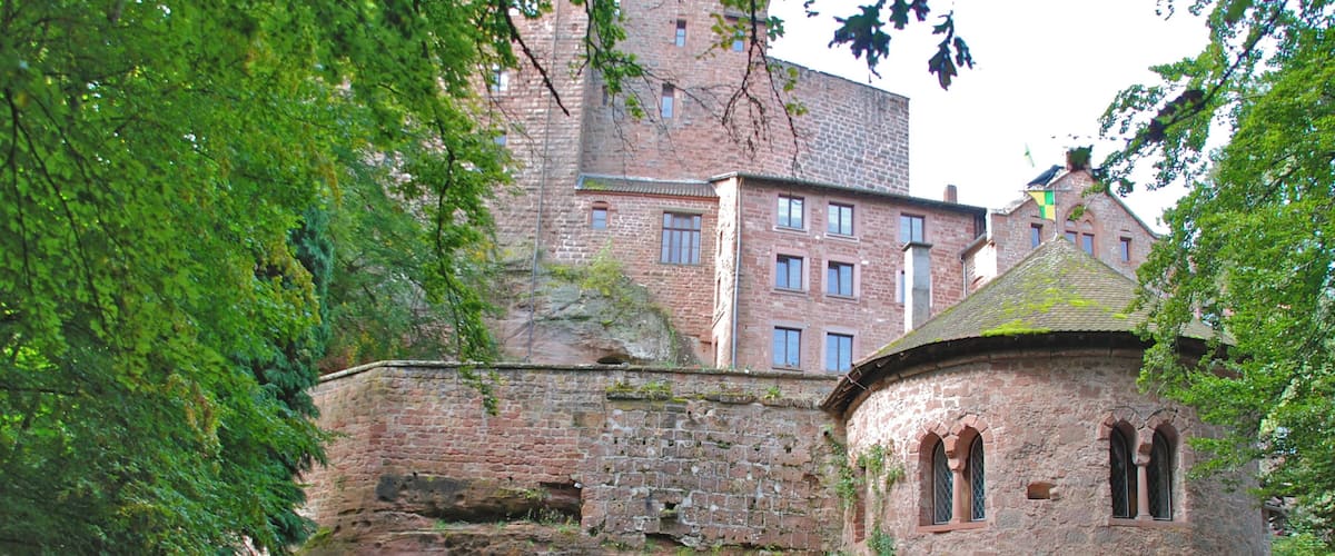 A historic stone building with arched windows and a moss-covered roof, surrounded by lush greenery and a pile of rocks.