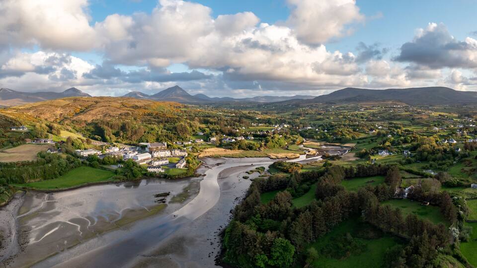 A river winding through a valley with houses and fields, surrounded by hills and mountains under a partly cloudy sky.