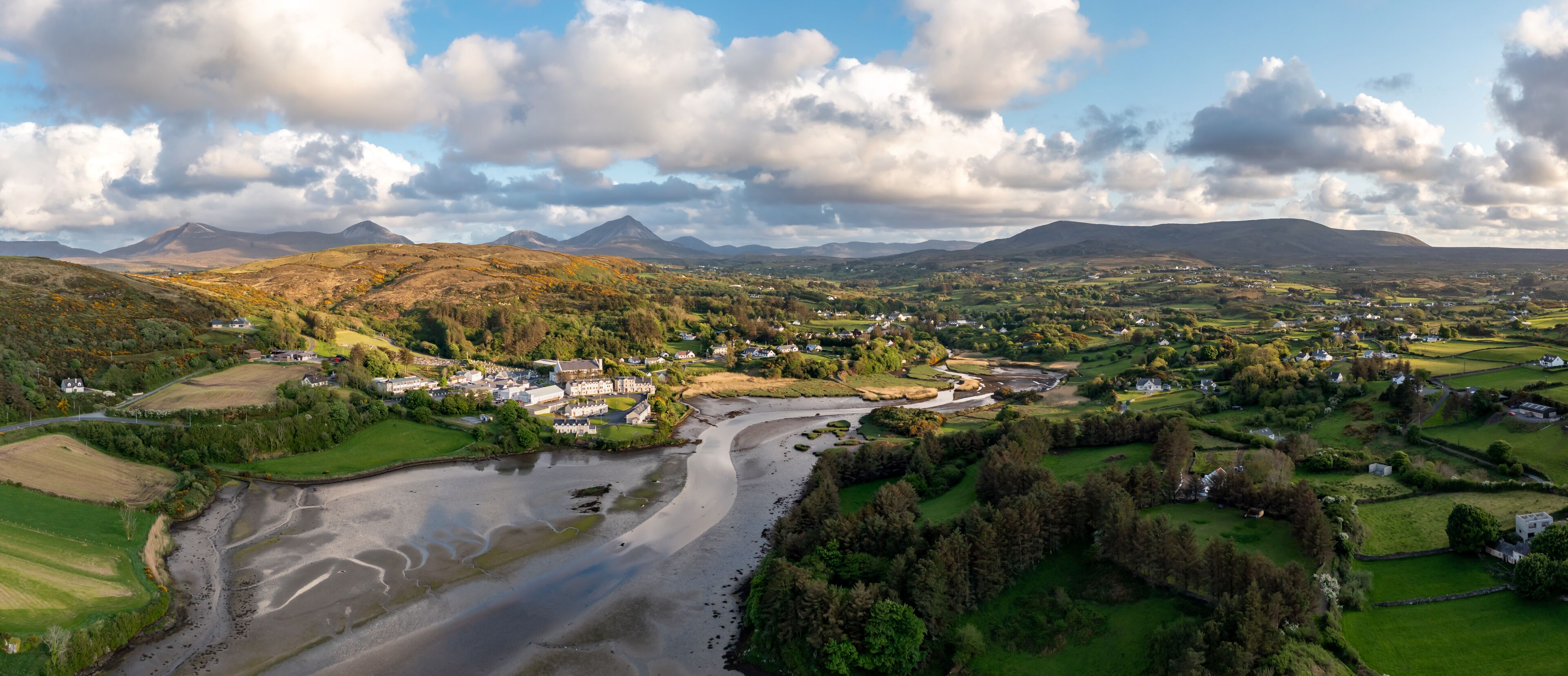 A river winding through a valley with houses and fields, surrounded by hills and mountains under a partly cloudy sky.