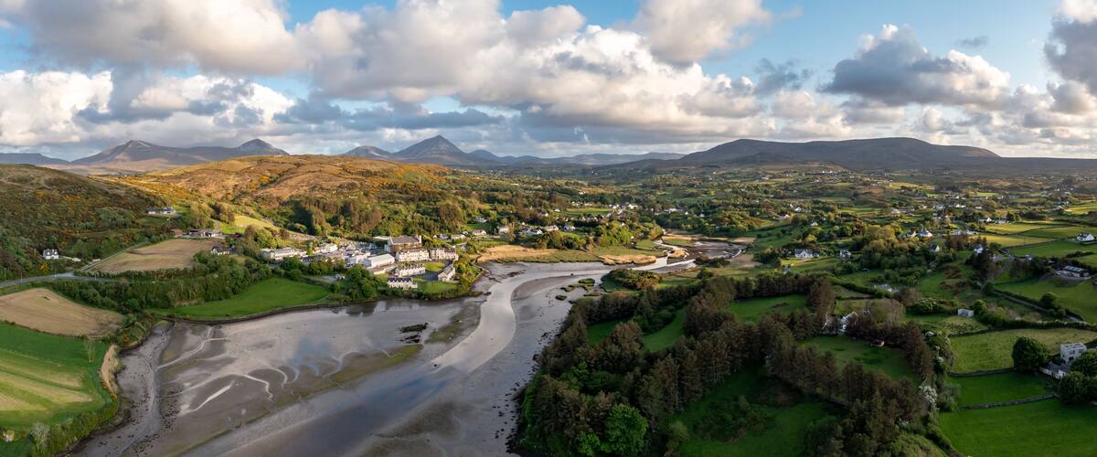 A river winding through a valley with houses and fields, surrounded by hills and mountains under a partly cloudy sky.