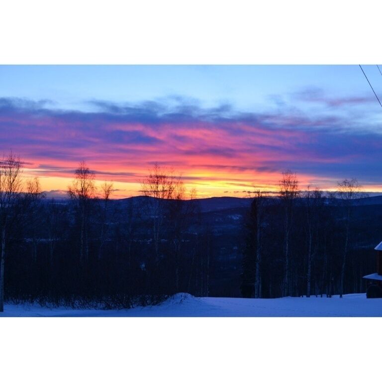 A sunset over a snowy landscape with bare trees and a distant cabin.