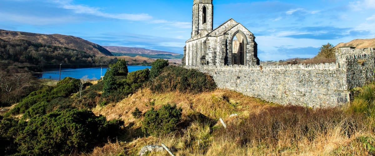 A historic stone church with a tall bell tower, surrounded by hills and a lake.