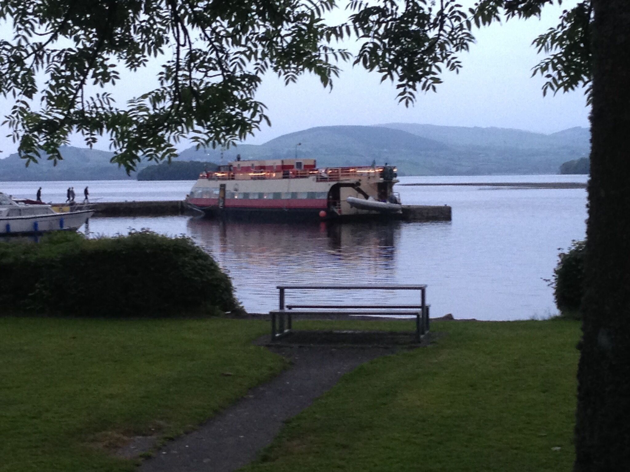 A river with a large boat docked at a pier, surrounded by hills and greenery.