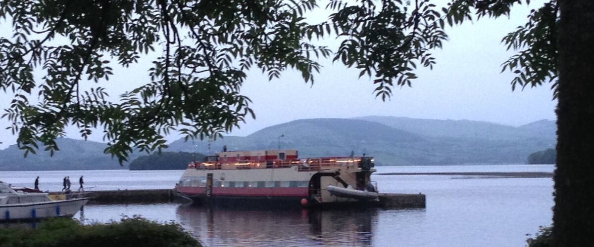 A river with a large boat docked at a pier, surrounded by hills and greenery.