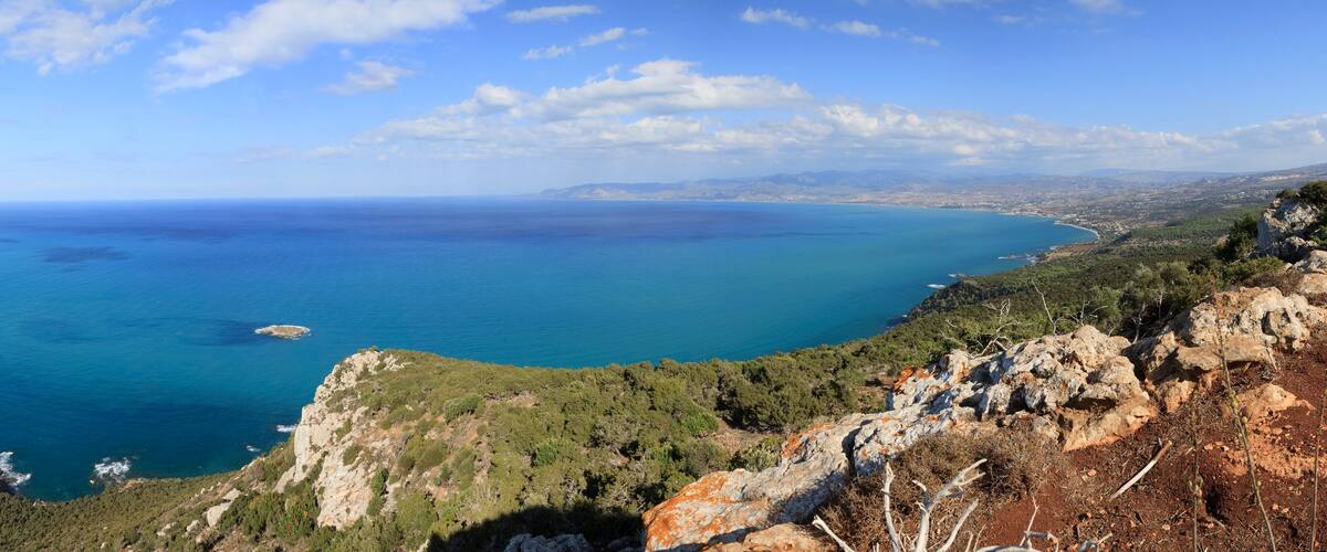 A coastal landscape with a clear blue sea, rocky cliffs, and dense greenery.