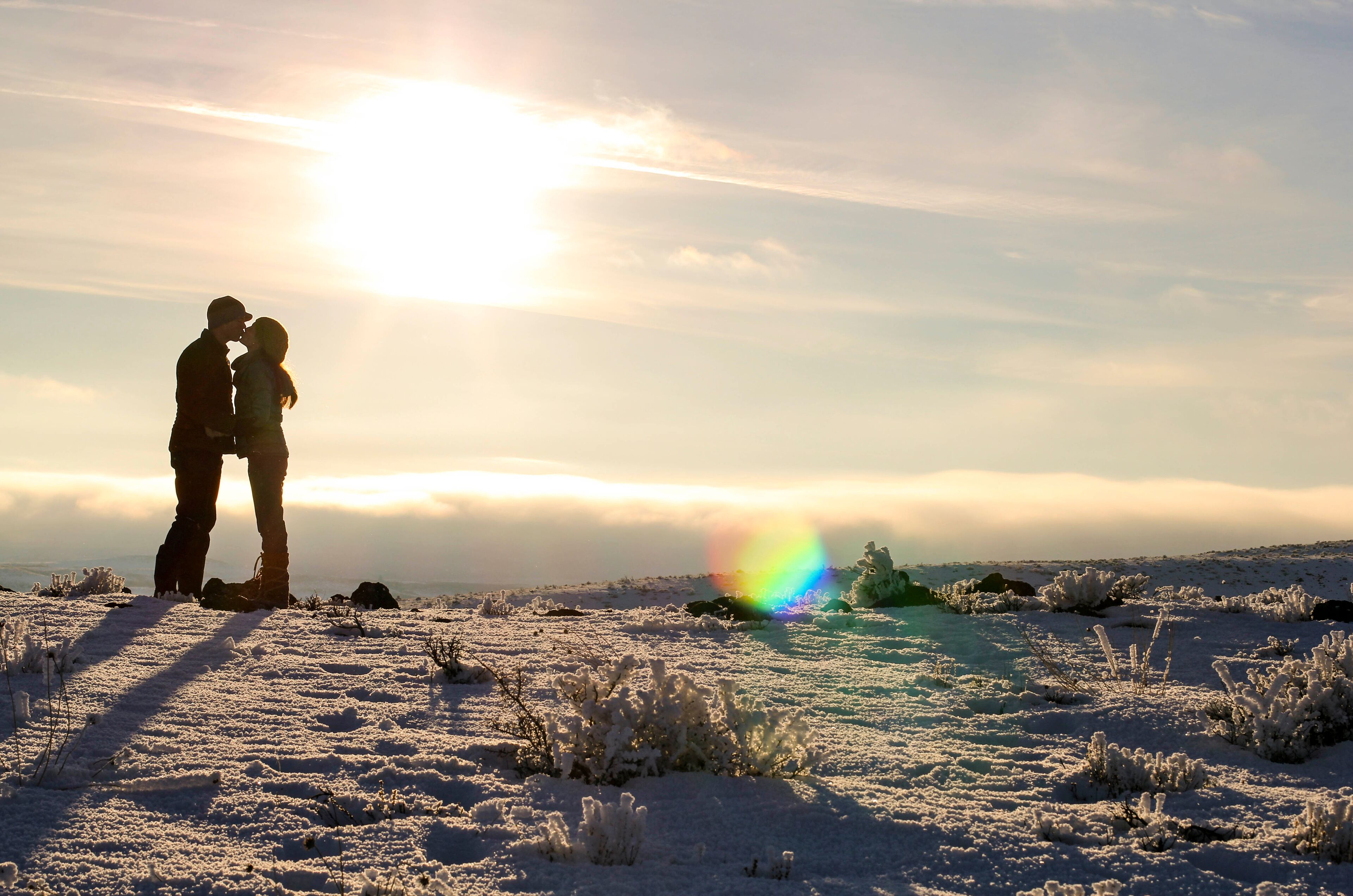 A couple embracing on a snow-covered landscape with a rainbow in the background.