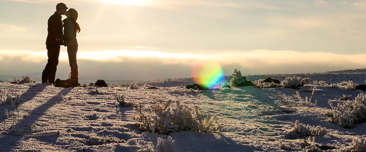 A couple embracing on a snow-covered landscape with a rainbow in the background.
