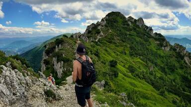 A hiker on a rocky mountain trail with lush green vegetation and a panoramic view of distant mountains.