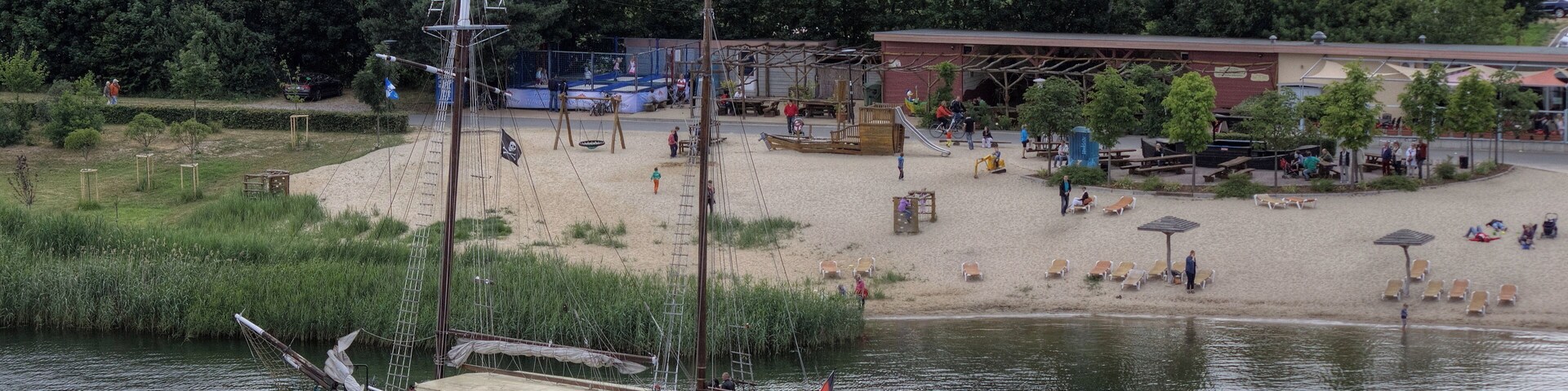 Un bote en un río tranquilo, una playa de arena con sombrillas y un área boscosa al fondo.