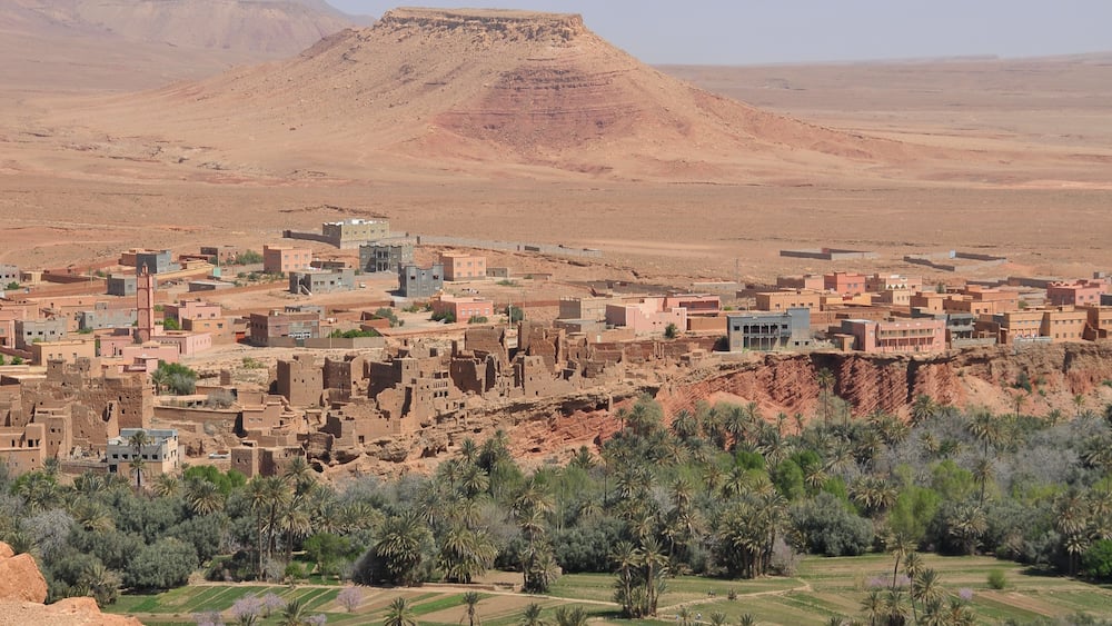 A desert landscape with a town built on a hillside, featuring traditional mud-brick buildings and palm trees.