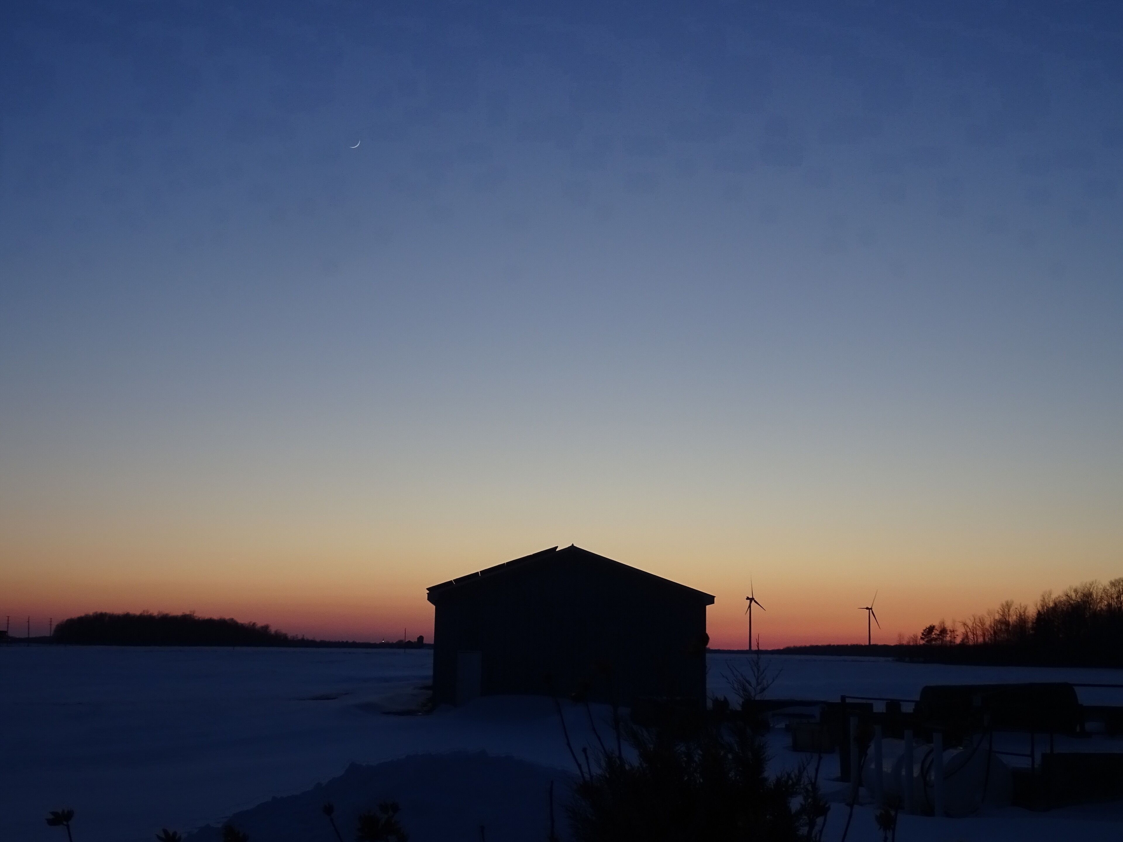 A rural landscape at dusk with a small building, snow-covered ground, and wind turbines.