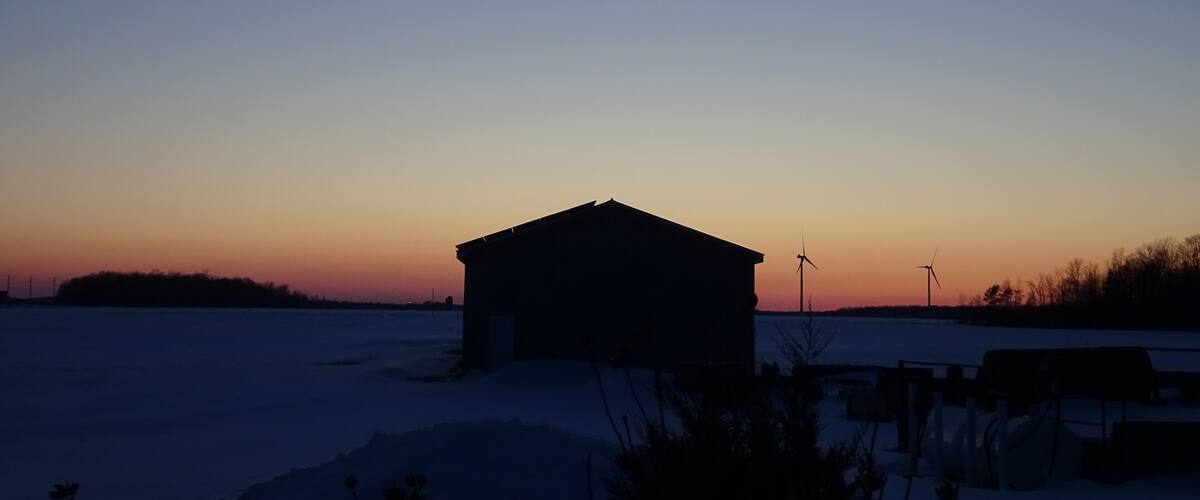 A rural landscape at dusk with a small building, snow-covered ground, and wind turbines.
