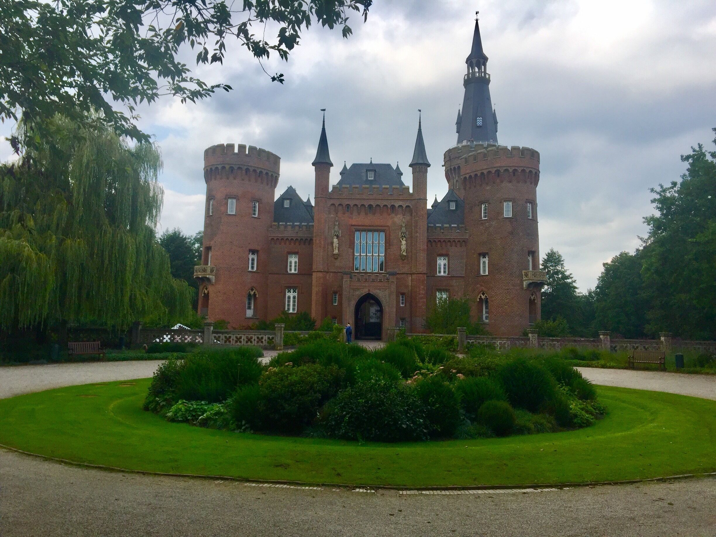 A large castle with multiple towers and a central entrance, surrounded by well-maintained gardens and a tree.