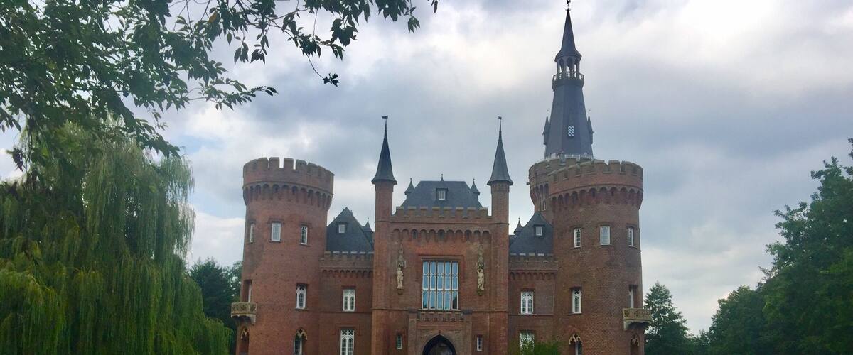 A large castle with multiple towers and a central entrance, surrounded by well-maintained gardens and a tree.