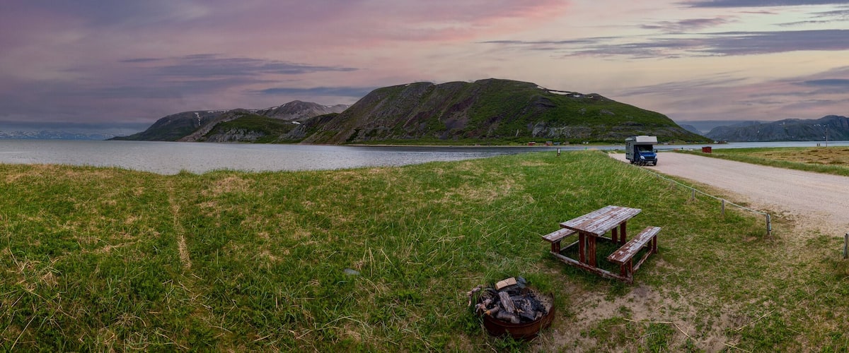 A gravel path leading to a picnic table and a truck parked near a body of water with mountains in the background.