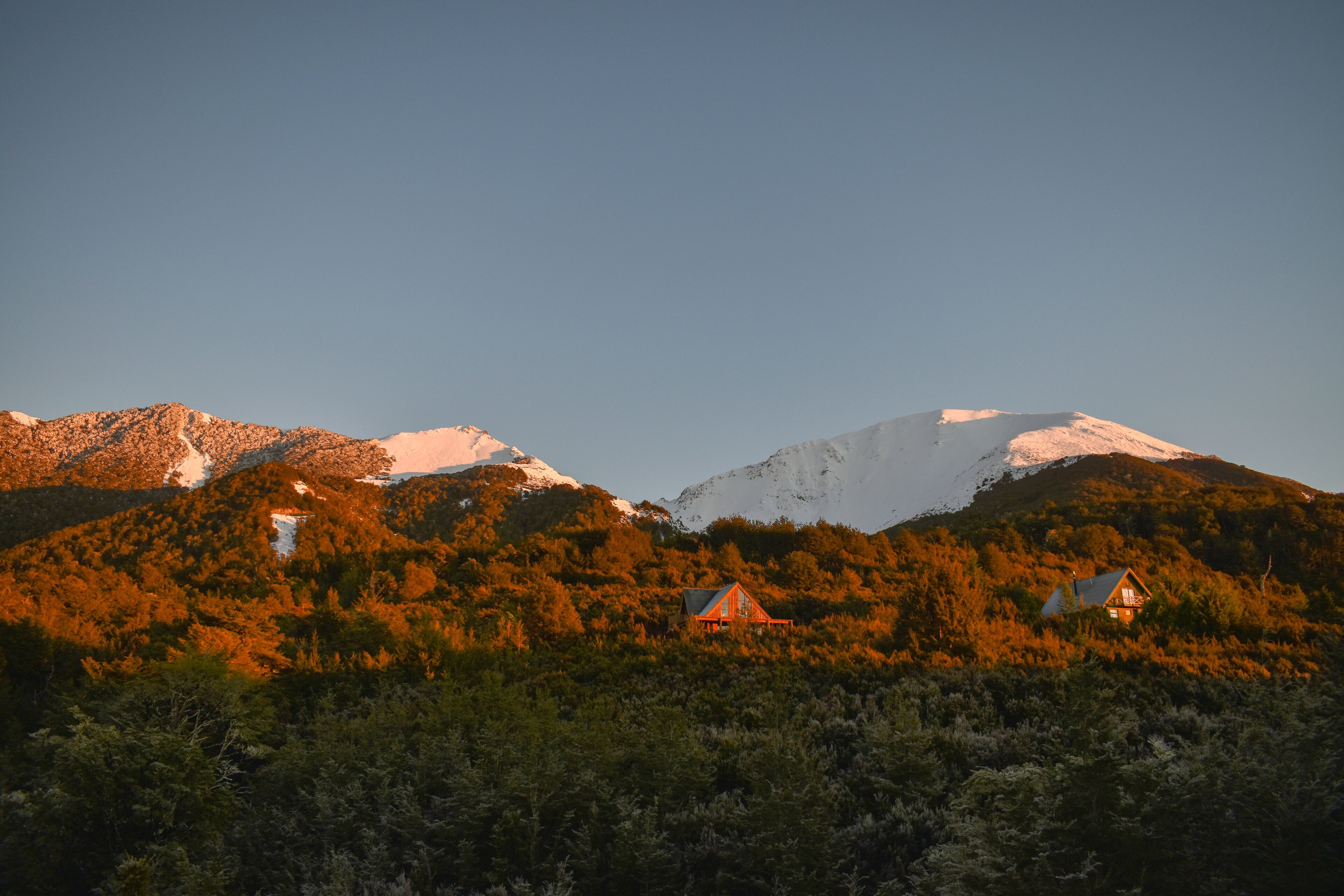 A mountain range with snow-capped peaks and a forested valley with two cabins.