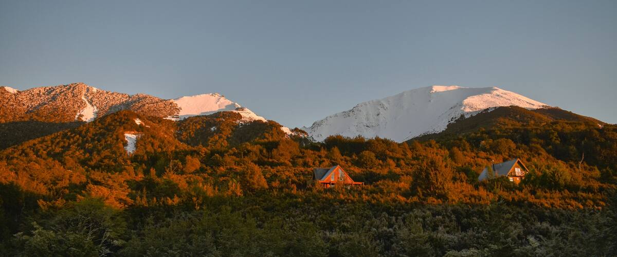 A mountain range with snow-capped peaks and a forested valley with two cabins.
