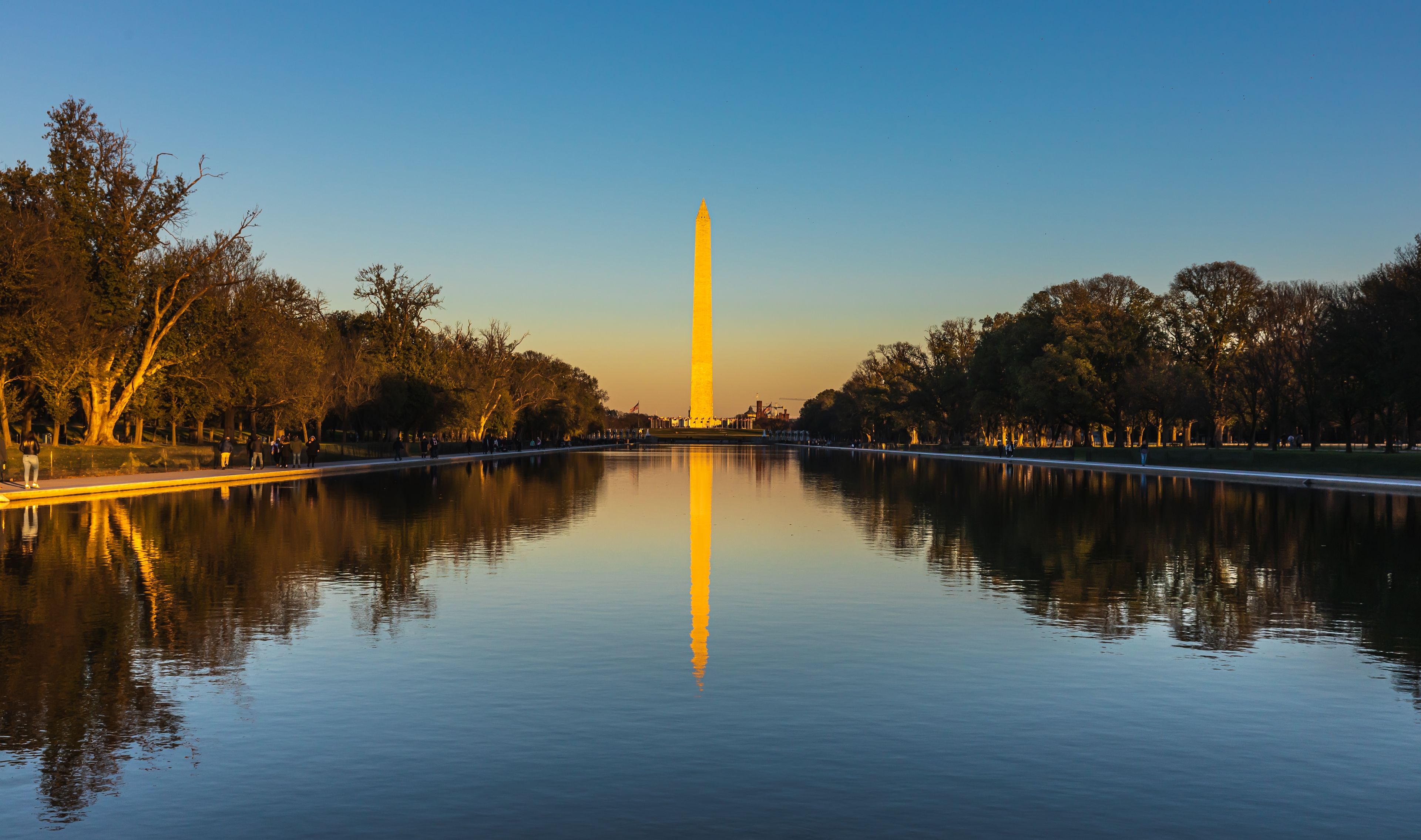 A calm river reflecting a tall monument, surrounded by trees and a clear sky.