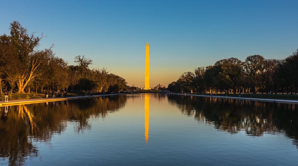 A calm river reflecting a tall monument, surrounded by trees and a clear sky.