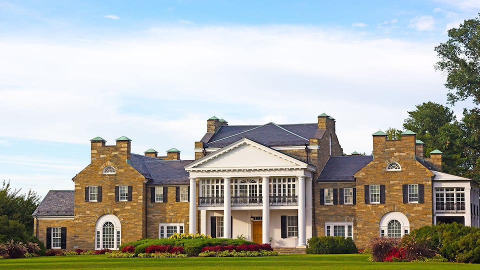 A large stone mansion with a central portico, surrounded by a well-manicured lawn and colorful flower beds.